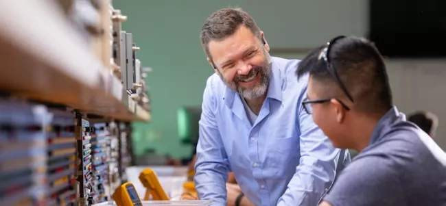 A man standing next to a student seated at a work station.