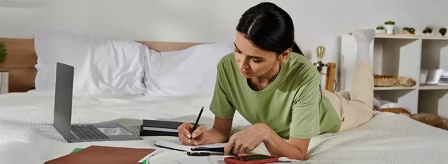 A woman laying on a bed with a laptop in front of her and writing in a notebook