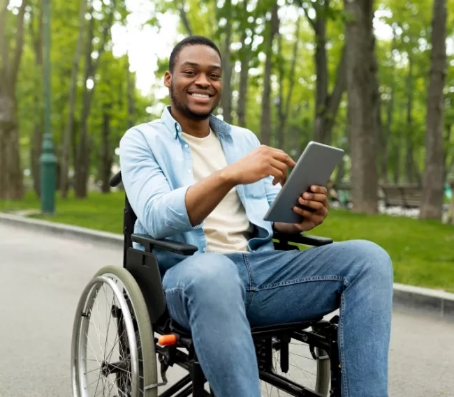 Man seated in a wheelchair outside on a pathway holding a tablet